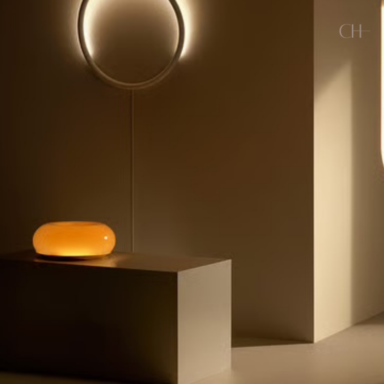 Bauhaus, a circular, donut-shaped, glass lamp, featured in orange, pictured on table, next to a modern circular wall light fixture, pictured in a minimally designed room.