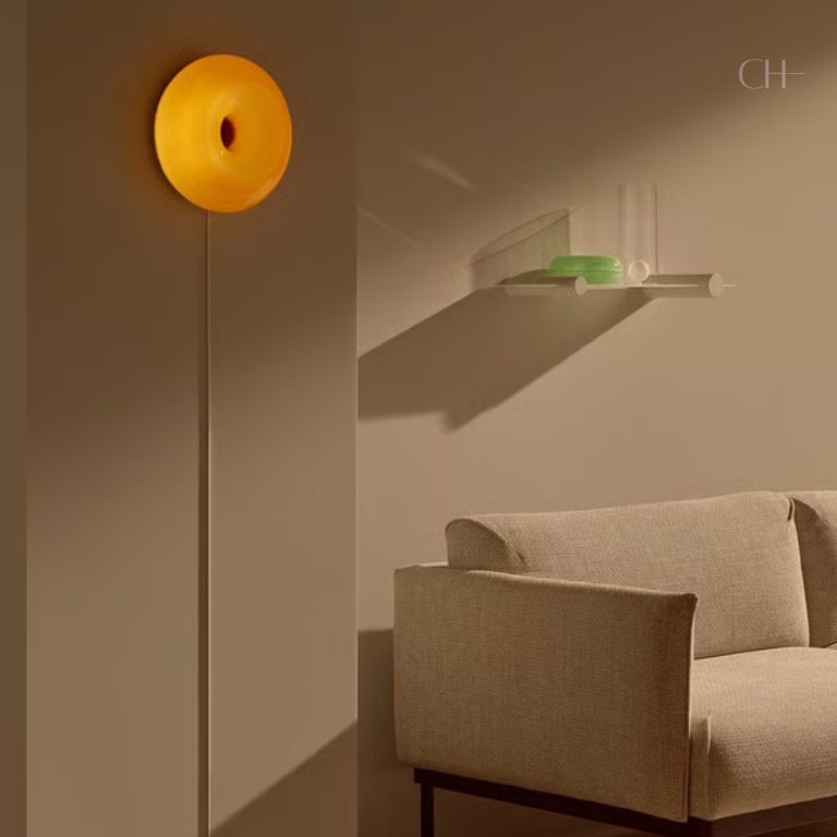 Bauhaus, a circular, donut-shaped, glass lamp, featured in orange, hung on a wall. Bauhaus is pictured next to a beige sofa, in an otherwise minimally designed room.