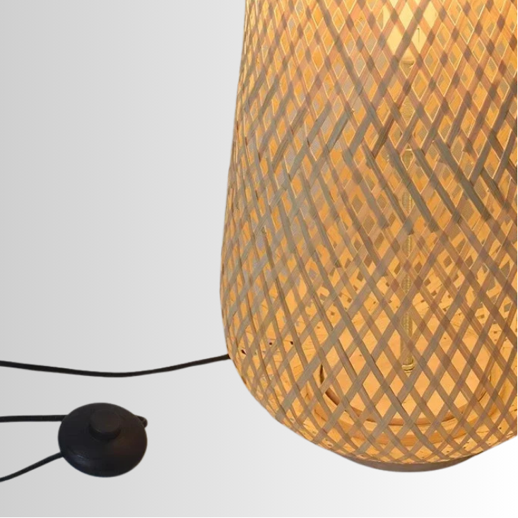 Solace, a cylindrical wicker floor lamp, in a natural light wheat color. The photo is slightly zoomed, focusing more on the base of the lamp with its button switch pictured. 