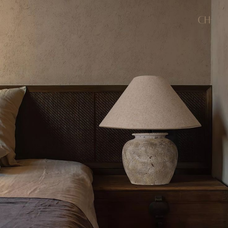 Kaia, a beige toned ceramic table lamp, pictured on a wooden bedside desk.