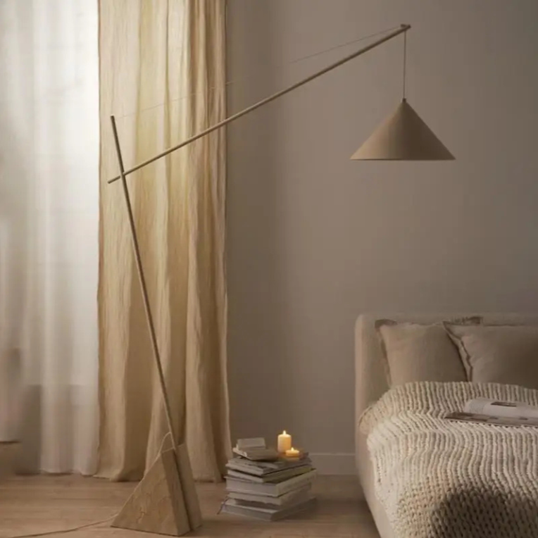Quentin, a sculptural and minimalist floor light with a beige hued iron body, and a triangular marble base. Pictured in a bedroom with ample natural light coming from the curtains, sat next to a stack of books.