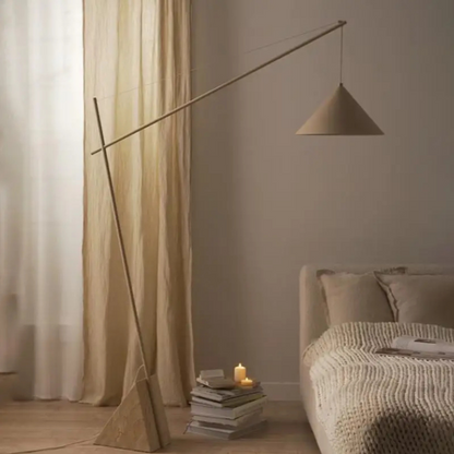 Quentin, a sculptural and minimalist floor light with a beige hued iron body, and a triangular marble base. Pictured in a bedroom with ample natural light coming from the curtains, sat next to a stack of books.