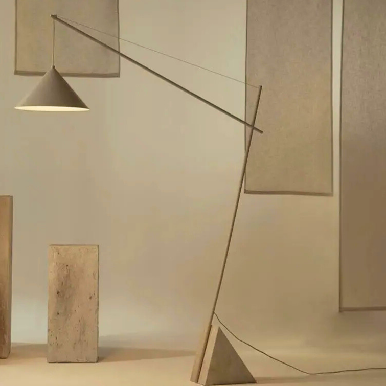 Quentin, a sculptural and minimalist floor light with a beige hued iron body, and a triangular marble base. Pictured in a bare room, with beige-toned minimalist paintings decorated on the walls.