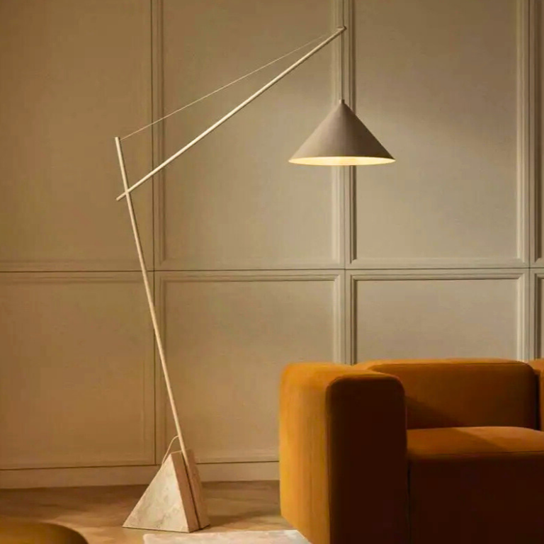 Quentin, a sculptural and minimalist floor light with a beige hued iron body, and a triangular marble base. Pictured in a room decorated with a burnt orange sofa.