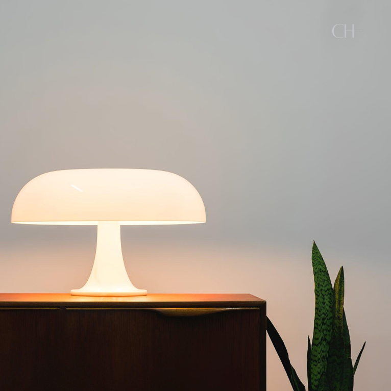 Giancarlo, an artistic, ivory colored modern table lamp with a silhouette resembling a mushroom, pictured on a desk next to a house plant.