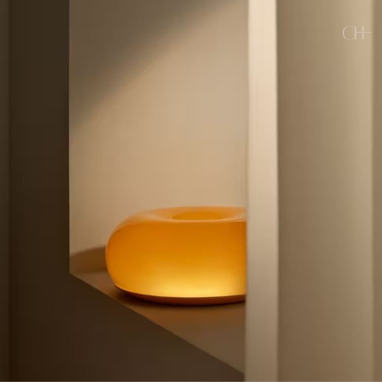 Bauhaus, a circular, donut-shaped, glass lamp, featured in orange, partially visible, pictured on window sill.