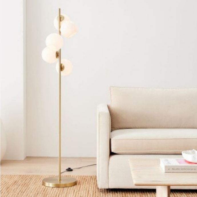 Galileo, a modern floor lamp with a gold-colored iron body and frosted glass bulbs, in a living room setting with a sofa and coffee table.