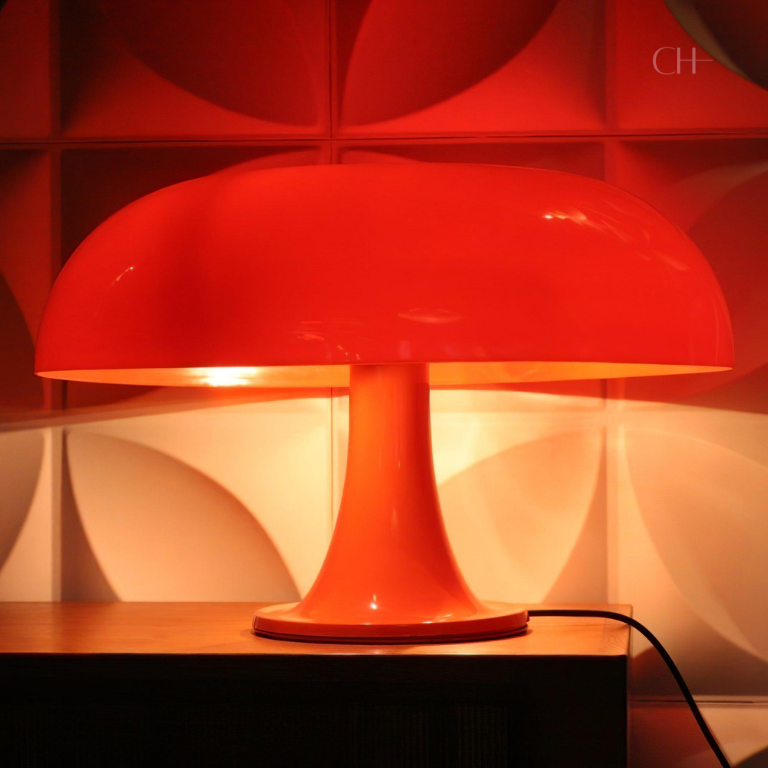Giancarlo, a modern orange table lamp with a silhouette resembling a mushroom, pictured on a desk as it emits an orange hue of light. 