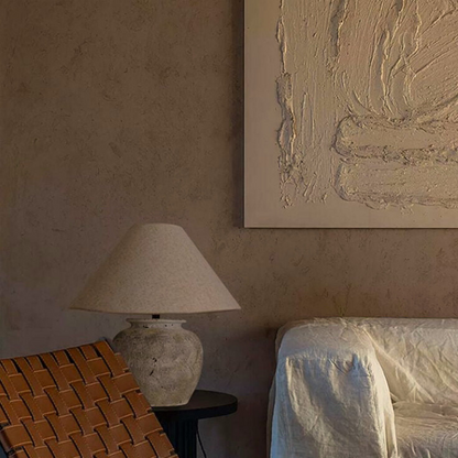 Kaia, a beige toned ceramic table lamp, pictured in an earth hued room, with a brown weave hair and a beige abstract painting on the wall.